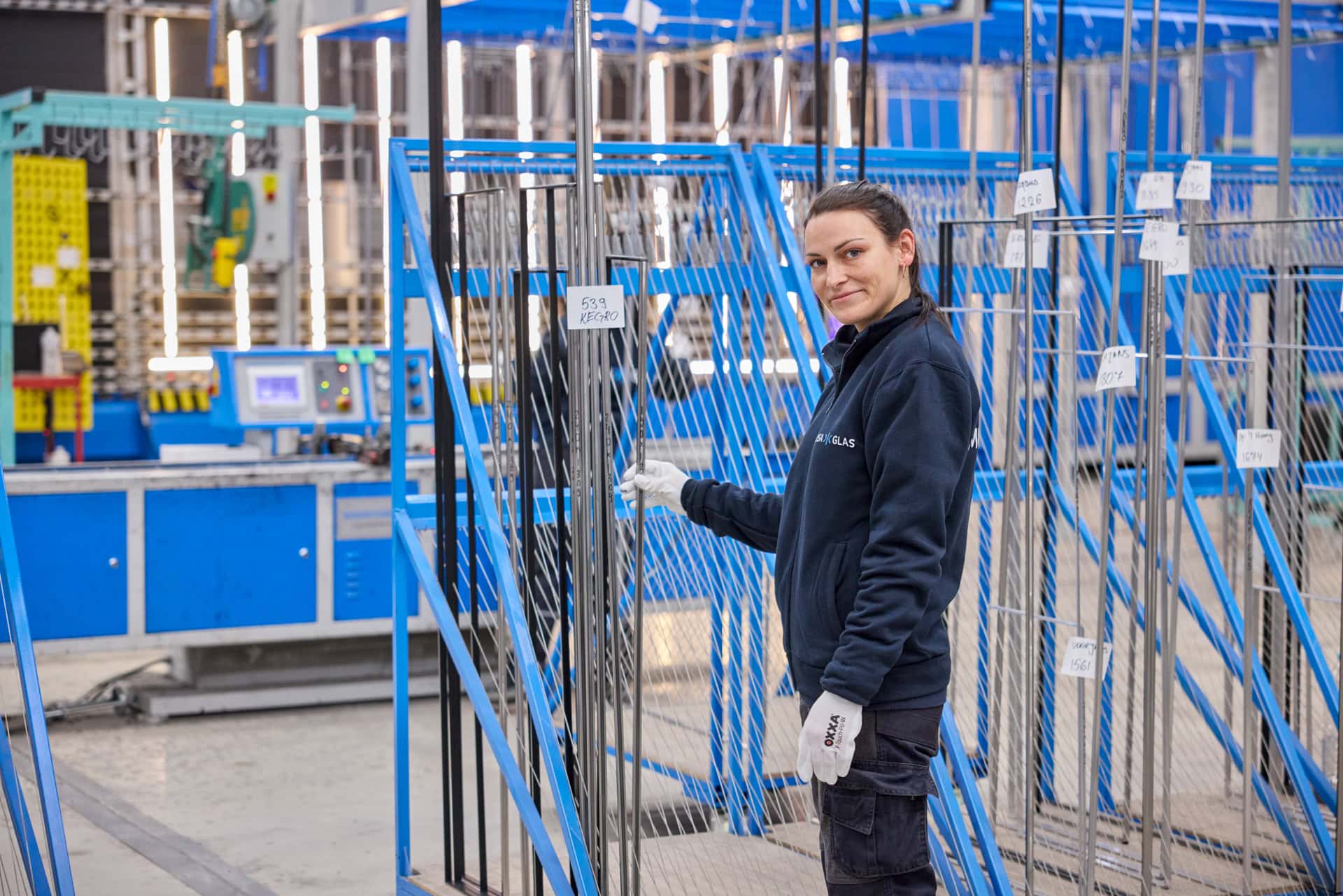 Een werknemer van Mosa Glas in een fabriekshal staat tussen spouw elementen die in blauwe metalen frames zijn geplaatst. De werknemer draagt een donkerblauwe jas en witte handschoenen, en houdt een van de spouw elementen vast. Op de achtergrond zijn meer machines en apparatuur te zien.