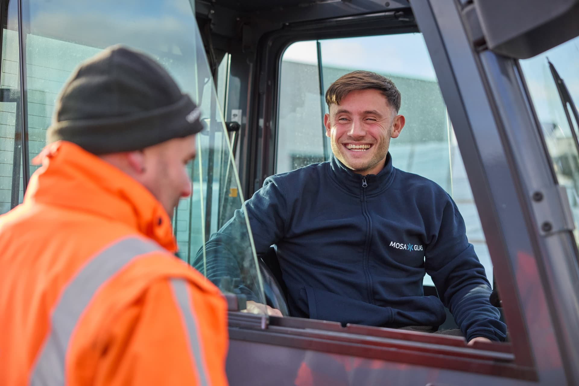 Twee personen in gesprek, waarvan één in een oranje veiligheidsjas en de ander in een cabine van een voertuig met het logo "Mosa Glas" op de trui