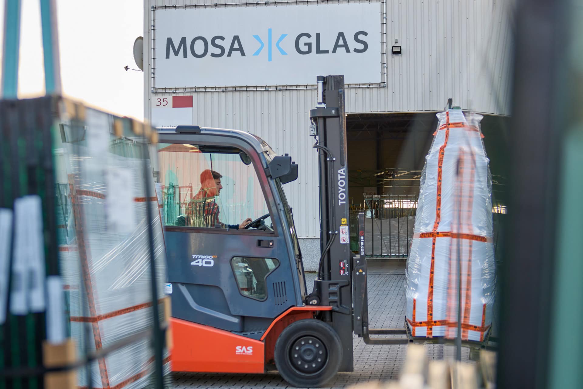 Een vorkheftruck bestuurd door een persoon in werkkleding, verplaatst een grote glazen plaat verpakt in beschermend materiaal. Op de achtergrond is het gebouw van Mosa Glas te zien met een groot bord waarop "Mosa Glas" staat