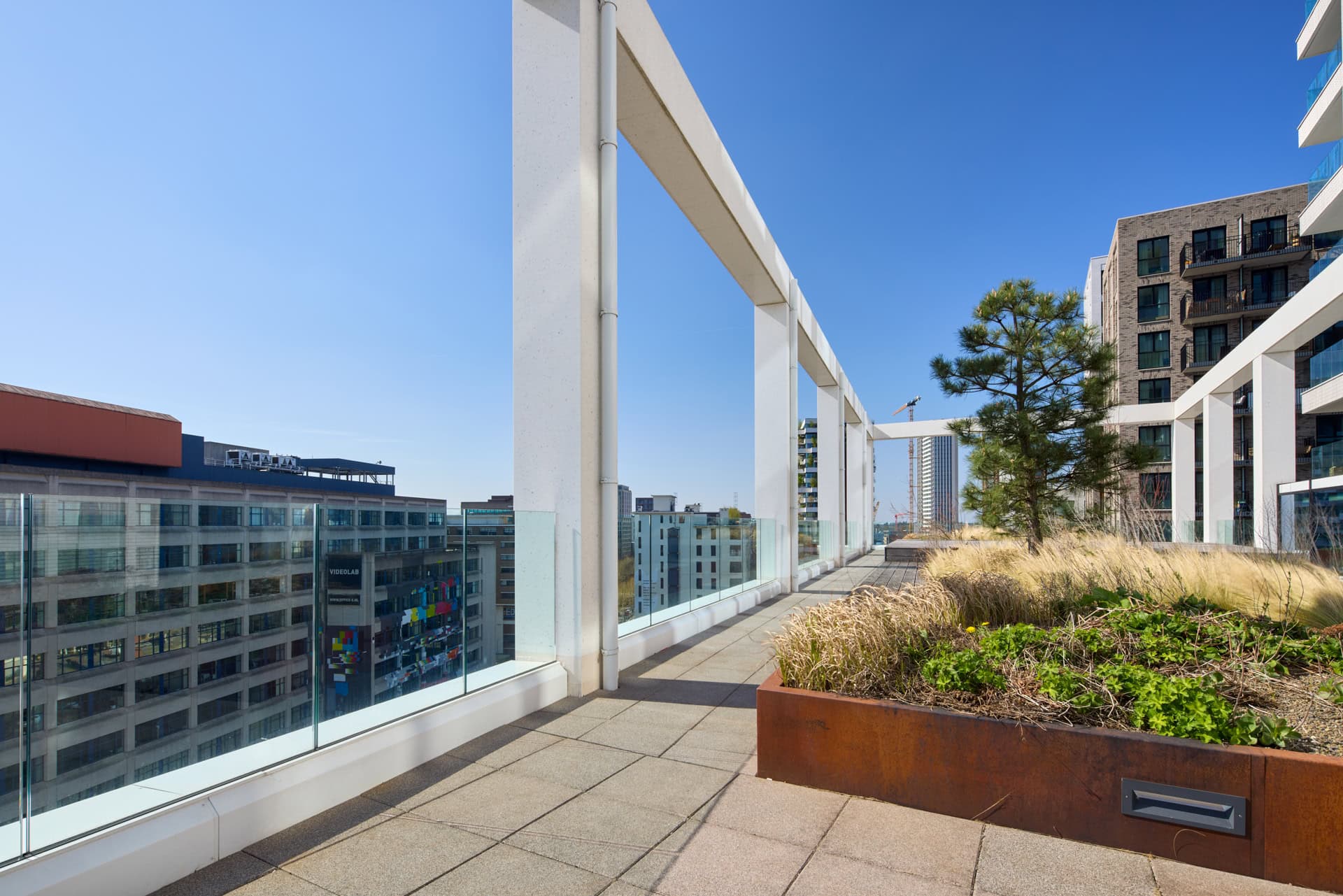 Een modern dakterras met enkel glas (gelaagd veiligheidsglas) balustrades, omgeven door hoge gebouwen en een blauwe lucht.