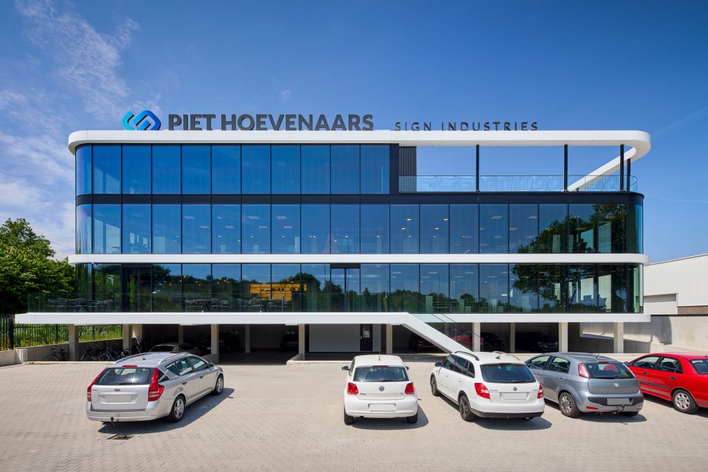 Modern kantoorgebouw van Piet Hoevenaars Sign Industries met glazen gevel en parkeerplaats op de voorgrond.