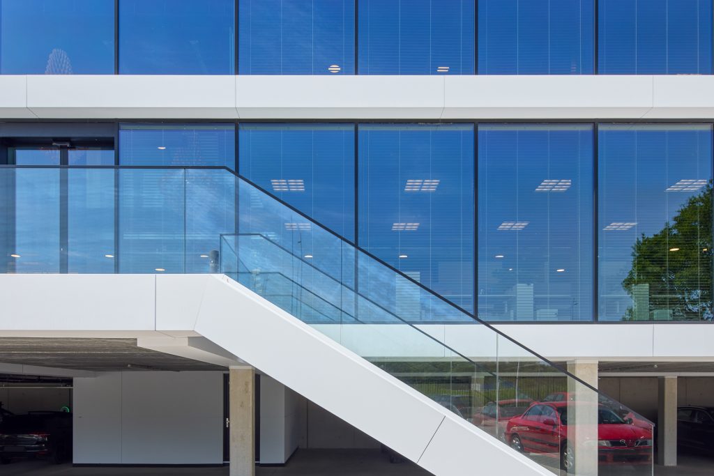 Modern gebouw met witte gevel, grote glazen ramen en een buitentrap met glazen balustrade.