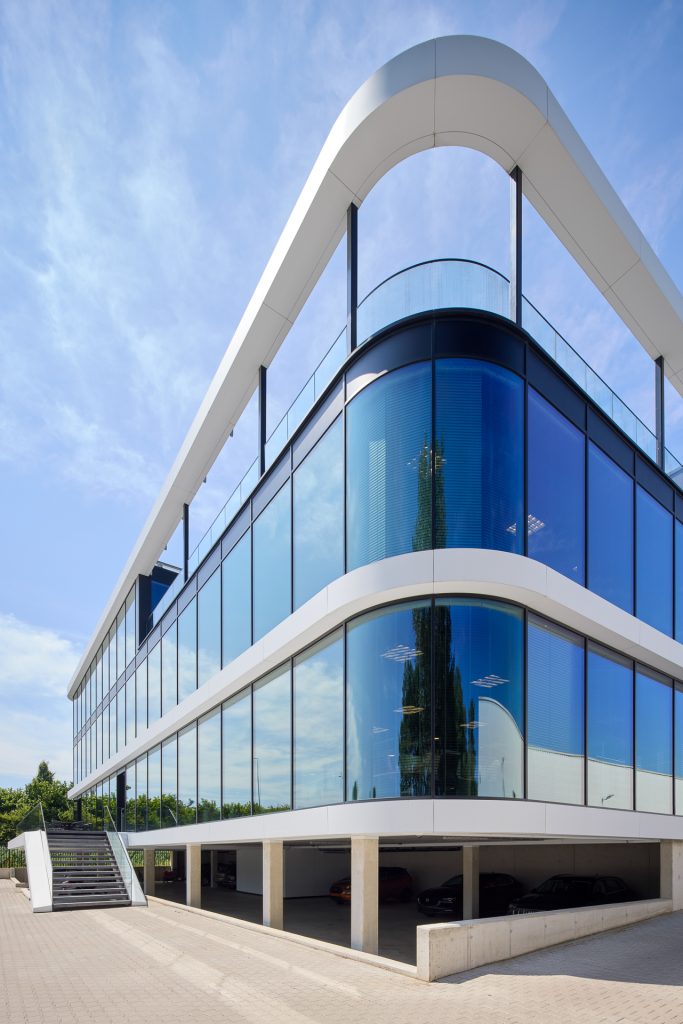 Modern gebouw met grote reflecterende ramen, witte structuur en parkeerruimte op de begane grond.