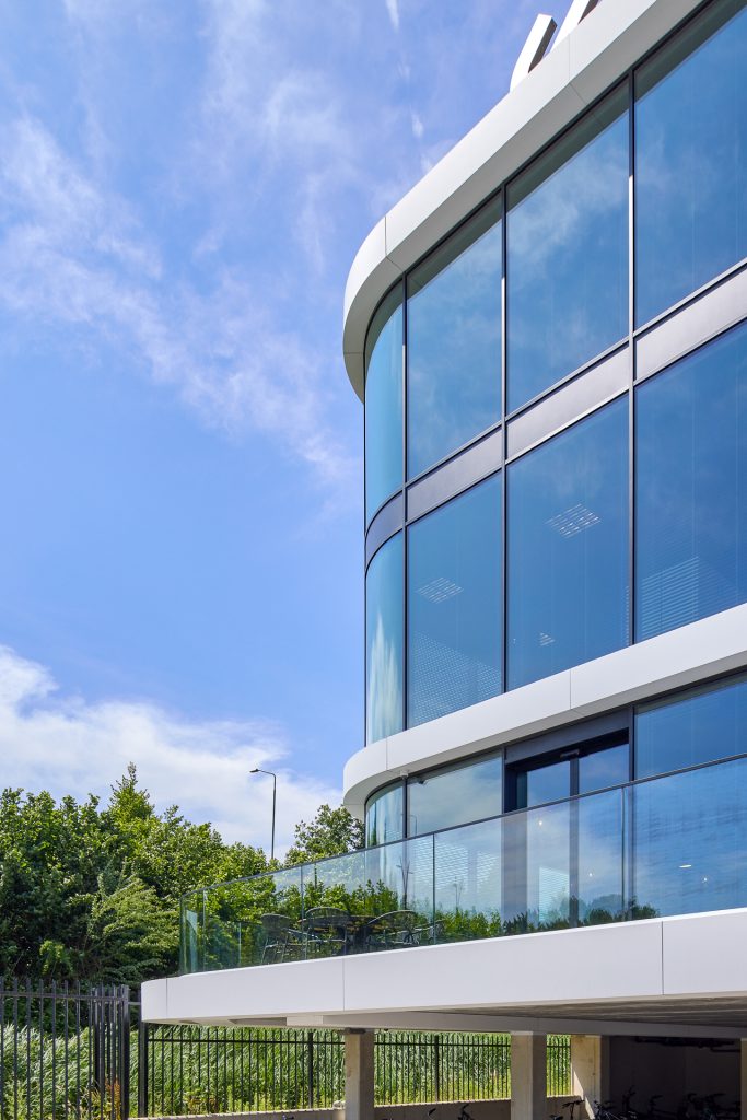 Modern gebouw met gebogen ontwerp, glazen ramen, balkon en fietsenstalling onder kolommen.