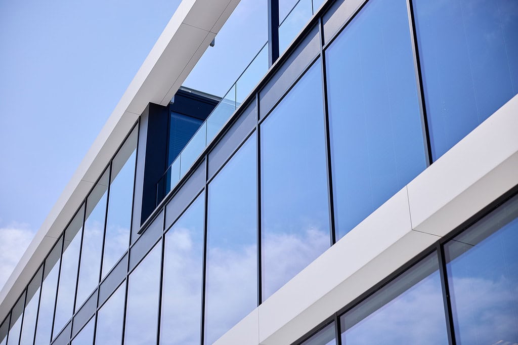 Modern gebouw met witte structuur en grote glazen ramen die de blauwe lucht en wolken reflecteren.