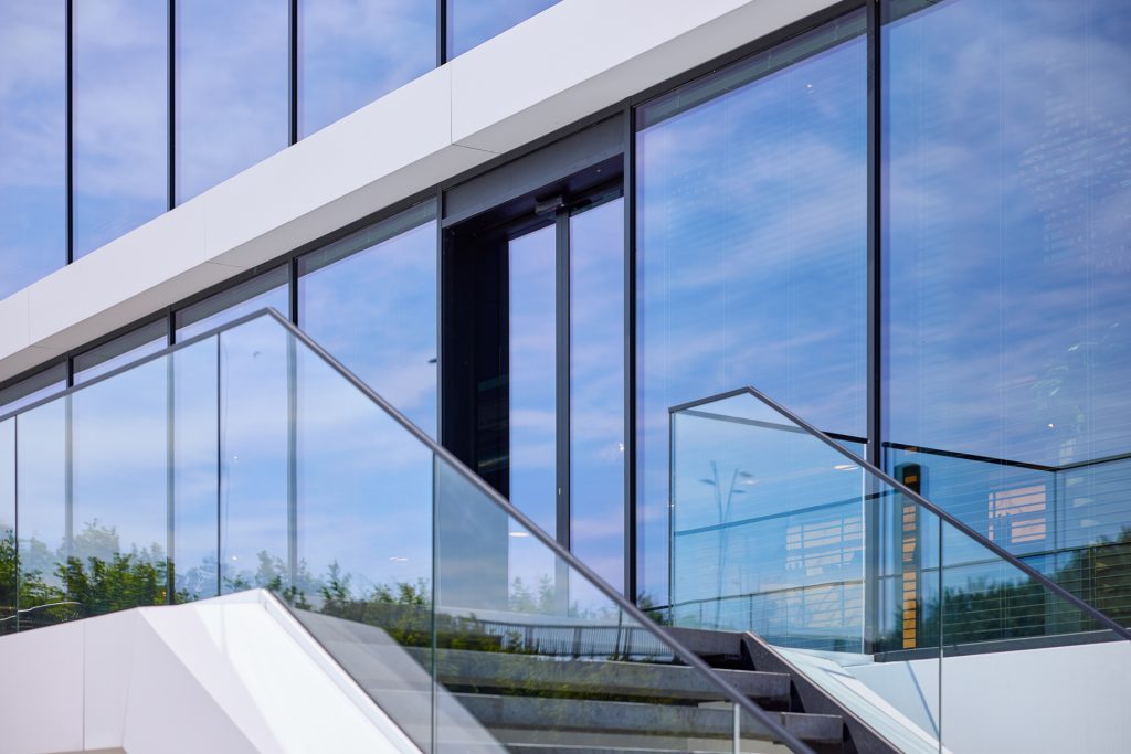 Moderne gevel met grote glazen ramen, glazen trapleuning en weerspiegeling van lucht en groen.